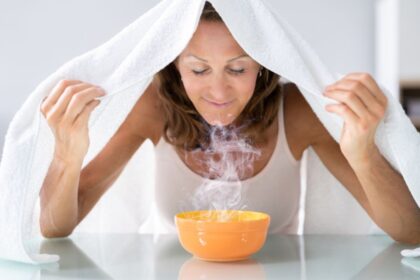 Egy nő gőzölést végez egy narancssárga tál fölött, törölköző alatt ülve.
