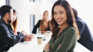 Egy nő mosolyogva ül egy tárgyalóasztalnál, háttérben másokkal.