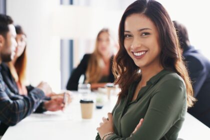 Egy nő mosolyogva ül egy tárgyalóasztalnál, háttérben másokkal.