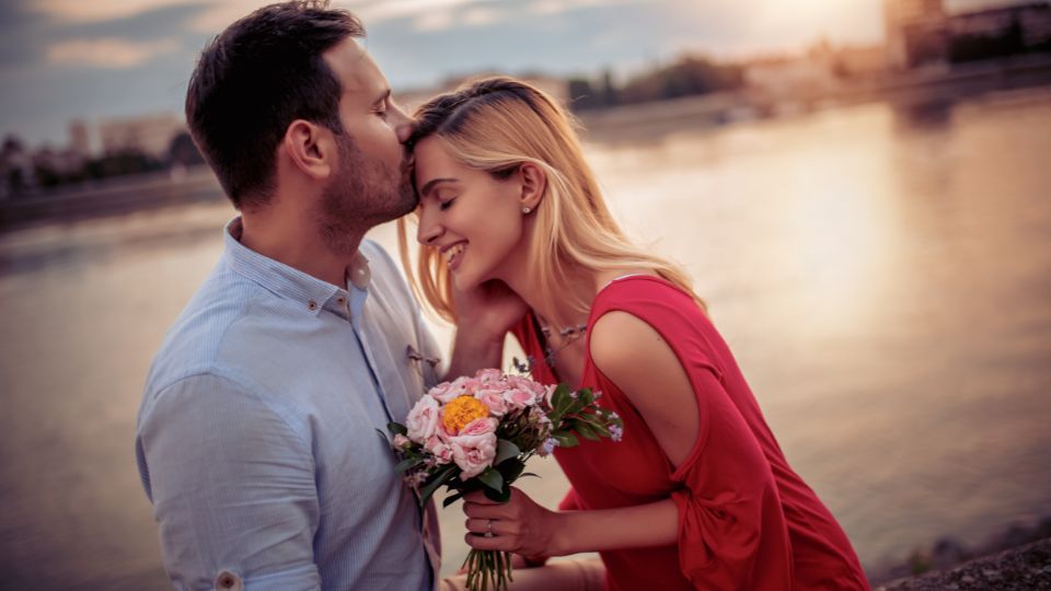 Egy férfi és nő romantikus pillanatot oszt meg a vízparton, virágcsokorral.
