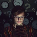 Fiatal férfi okostelefonon válaszra vár, órák és kérdőjelek között.