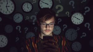 Fiatal férfi okostelefonon válaszra vár, órák és kérdőjelek között.