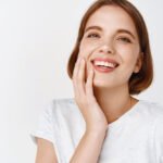 Egy fiatal nő mosolyogva érinti meg arcát, egészséges, fehér fogaival.